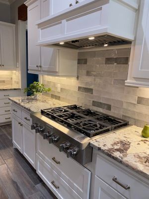 A kitchen stove with white cabinets, light stone countertops, and a gray-toned tile backsplash under a white range hood.