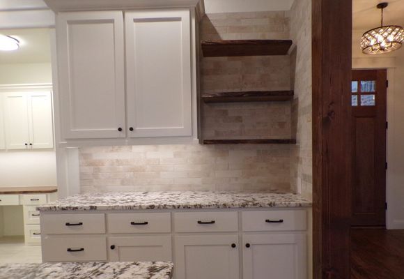 White kitchen cabinets with granite countertops, a stone backsplash, and floating wood shelves next to a dark wood doorway.
