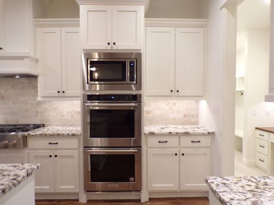 White kitchen cabinets with stainless steel double ovens and a microwave set between stone countertops and a backsplash.