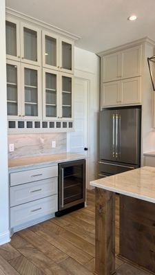 A kitchen featuring light gray cabinetry with glass upper doors, a stainless wine cooler, and a stainless refrigerator.