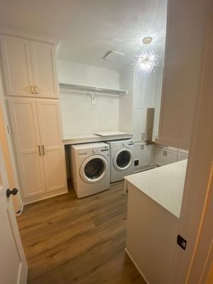 A bright laundry room with white cabinets, a washer, a dryer, and wood-look flooring under a modern light fixture.