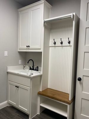 A white mudroom cabinet featuring a sink with a black faucet, three coat hooks, and a wooden bench with shoe storage below.