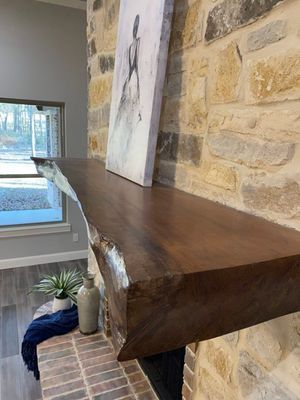 A live-edge dark wood mantelpiece installed on a rustic stone fireplace with a framed print leaning against the wall.