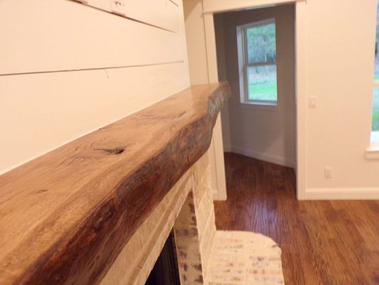 A natural-edge wood mantel sits above a light stone fireplace, with white shiplap walls and a nearby doorway in view.