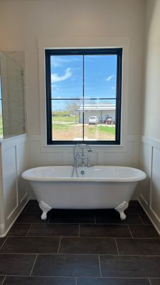 A white clawfoot bathtub centered in front of a window with black trim, set against white wainscoting and dark floor tiles.