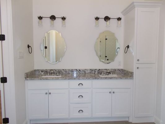 A double-vanity bathroom with white cabinets, granite countertop, two oval mirrors, wall lights, and a tall storage unit.
