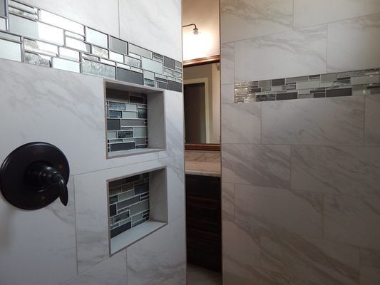 A bathroom shower interior featuring large white marble-look tiles, two recessed wall niches, and a decorative mosaic strip.