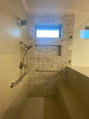 A modern walk-in shower with light-colored brick walls, a built-in bench, a window, a handheld showerhead, and grab bars.