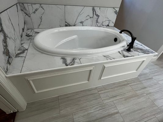 Oval white bathtub set in a marble-look tiled deck with white wainscoting and light wood-look floor tiles.