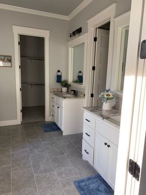 A brightly lit bathroom with white vanity cabinets, grey tile floors, two mirrors, and an open doorway to a walk-in closet.