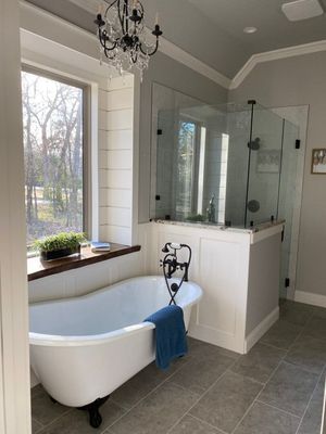 A bathroom with a freestanding white tub, a walk-in glass shower, a chandelier, and a large window with a wood ledge.