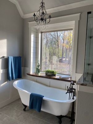 A freestanding white clawfoot tub sits below a window with a wood ledge in a bathroom with a hanging chandelier.