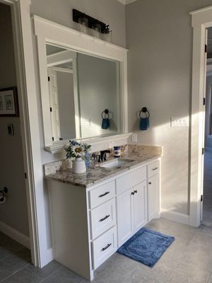 A bathroom vanity with white cabinets, a granite countertop, a large framed mirror, and a blue rug on a tiled floor.