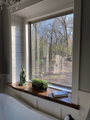 A live-edge wood window sill with a small green plant, a decorative heron, and a book, overlooking a wooded forest.