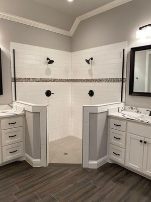 A walk-in corner shower with white subway tile and black fixtures, situated between two white bathroom vanities.