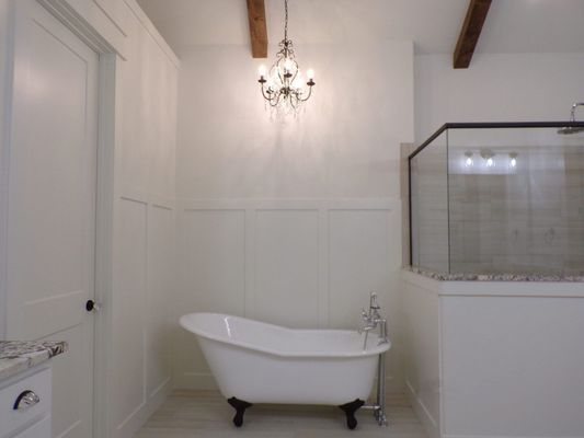 A white claw-foot bathtub under a crystal chandelier in a bathroom with white paneling and exposed wood ceiling beams.