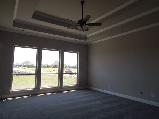 An empty room with gray carpet, neutral walls, a tray ceiling with a fan, and three large windows overlooking a field.