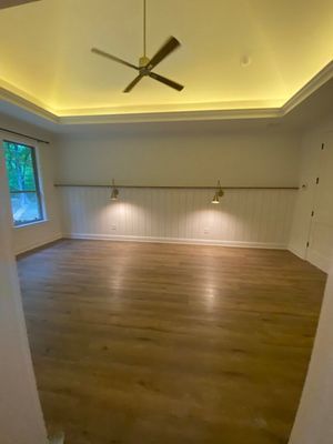 An empty room with light wood floors, white vertical wall paneling, two wall sconces, a tray ceiling, and a ceiling fan.