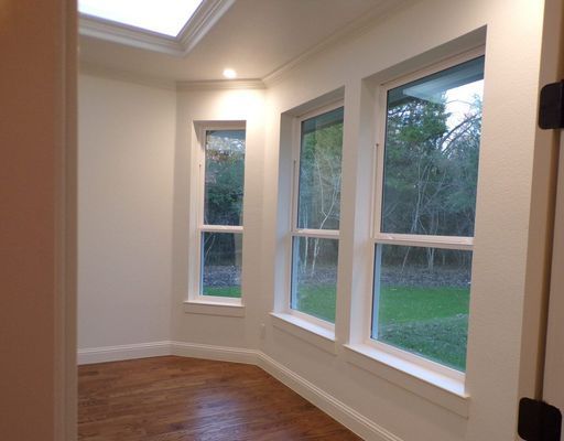 A sunlit room with hardwood floors, white walls, and a large bay window overlooking trees and a green lawn.