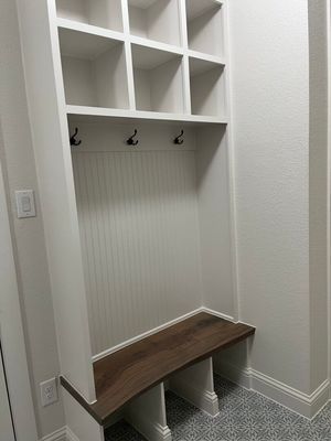 A white mudroom built-in unit with top cubbies, three black wall hooks, and a dark wood bench seat.
