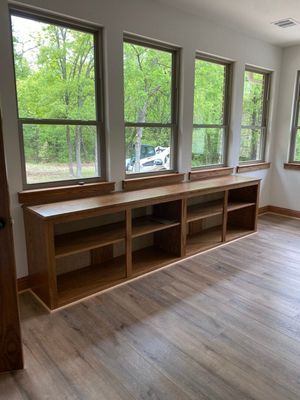 Low wooden bookshelves built beneath a row of four windows overlooking a green, wooded yard in a room with wood flooring.