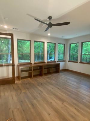 A bright, empty room with light wood floors, a dark ceiling fan, and large windows looking out onto a green forest.