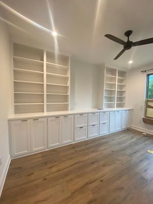 A room with white built-in shelving and cabinets lining a wall, featuring light wood floors and a ceiling fan.