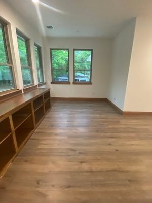 A room with light wood flooring, a long wooden built-in bookshelf along the wall, and three large windows overlooking trees.