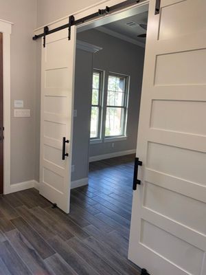 Two white, five-panel sliding barn doors mounted on a black rail, separating two rooms with gray walls and wood-look floors.