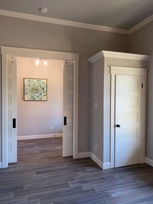 A room with gray wood-look floors, a wide entryway showing a succulent art piece, and a single white door with trim.
