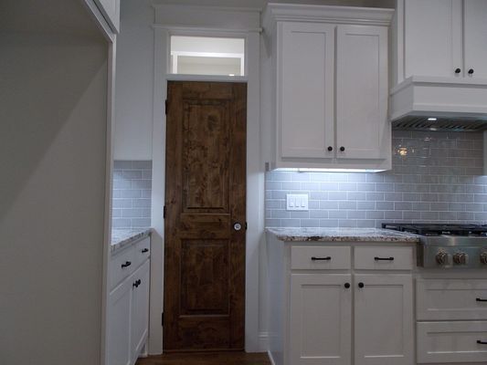 A white kitchen with gray subway tile backsplash, white cabinets, and a tall, dark wooden door with a glass transom window.