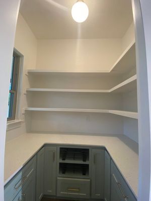 A walk-in pantry featuring sage green lower cabinets, white countertops, and three levels of L-shaped white wall shelving.