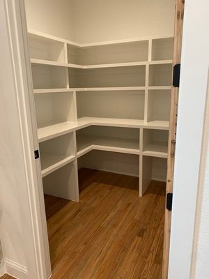 A walk-in pantry with white L-shaped shelves and wood-look flooring, viewed through a doorway.