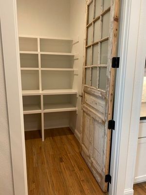 A pantry with white shelves and light wood flooring, accessed by a rustic, distressed-wood glass panel door.