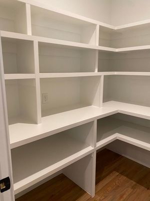 Empty walk-in pantry with white, custom-built wooden shelving units and light wood flooring.