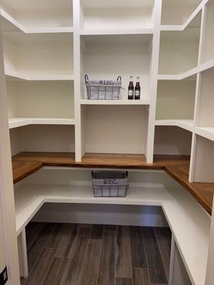 A walk-in pantry with white shelves, wood-toned counters, and two wire baskets on a dark wood-look tiled floor.