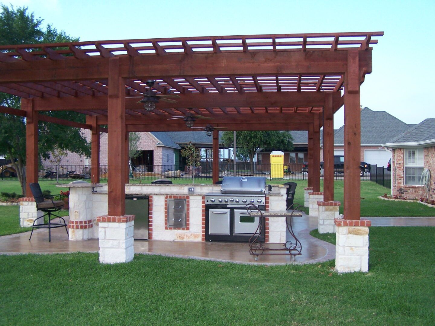 Outdoor wooden pergola over a stone-built kitchen island with a grill and stainless steel appliances in a backyard.