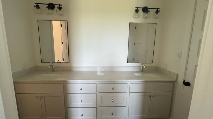 A double bathroom vanity with two rectangular mirrors, wall lights, and white cabinets against a plain white wall.
