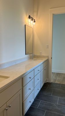 A bathroom vanity with white cabinets, a long countertop, dual sinks, a mirror, wall lights, and dark tiled flooring.