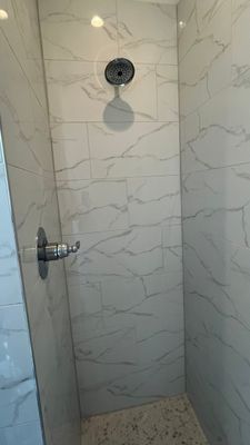 A walk-in shower with white marbled wall tiles, a metallic shower head, and a chrome valve handle.