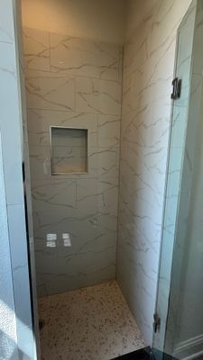 A shower with light marble-patterned wall tiles, a built-in recessed shelf, and small, neutral-colored floor tiles.
