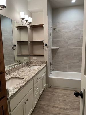 A modern bathroom featuring a double vanity with granite countertops, wood shelving, and a tiled shower-tub combo.