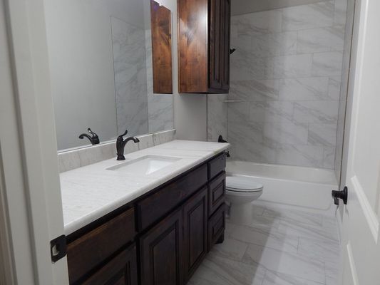A modern bathroom features a dark wood vanity, white countertop, marble-look tiled shower, and a toilet.