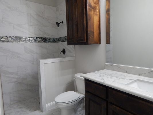 A bathroom with a tiled walk-in shower, a toilet, and a white marble-top vanity with dark wood cabinets.