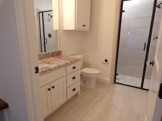 A bright bathroom with white cabinets, a light stone countertop, a toilet, and a glass-enclosed shower with tiled walls.
