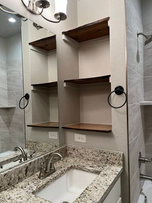 A bathroom vanity with a granite countertop, a sink, and wooden shelves built into the wall next to a mirror.