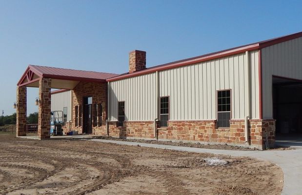 A single-story metal building with a stone facade, stone entrance columns, and a reddish metal roof under a blue sky.