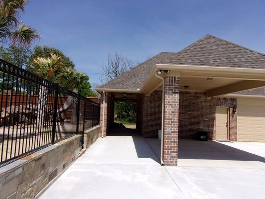 A brick house with a covered patio and concrete driveway, beside a black metal fence on a stone retaining wall.