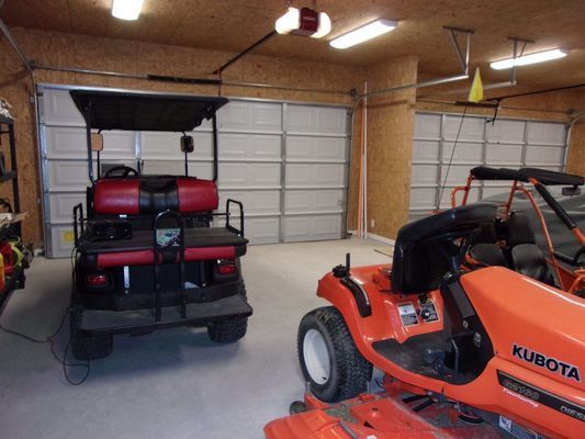 A golf cart with red and black seats parked next to an orange Kubota riding mower inside a garage with wood walls.