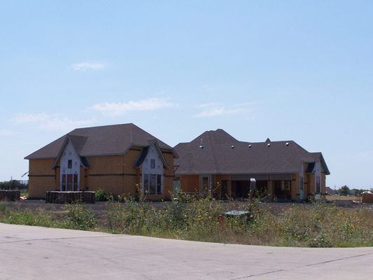 Two unfinished houses with brown roofs and wooden walls stand side-by-side in a field under a clear blue sky.